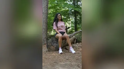 Smiling woman in pink graphic t-shirt and black shorts sitting on a log in a lush green forest.
