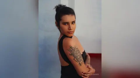 Tattooed woman in a black sleeveless top posing with a confident expression against a minimalist background.