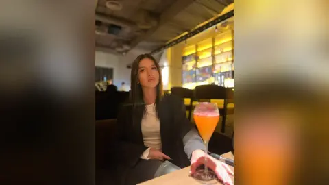JadaJosey sits at a restaurant table with long brown hair, wearing a black blazer over a white top, next to a glass of orange cocktail.
