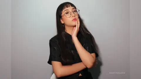 Stylish young woman with glasses and long dark hair wearing a black shirt in a minimalist studio setting.