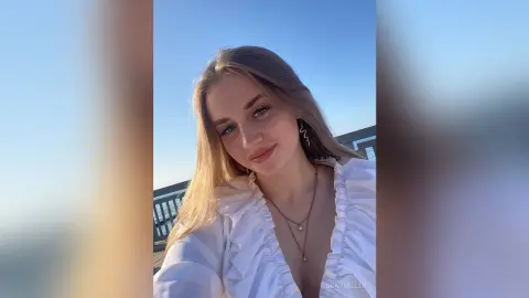 Smiling young woman in a white ruffled blouse posing outdoors against a clear blue sky.