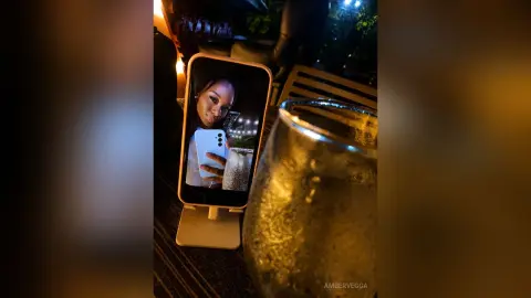 Smiling woman taking a selfie on her phone at a dimly lit outdoor evening lounge with a refreshing drink.