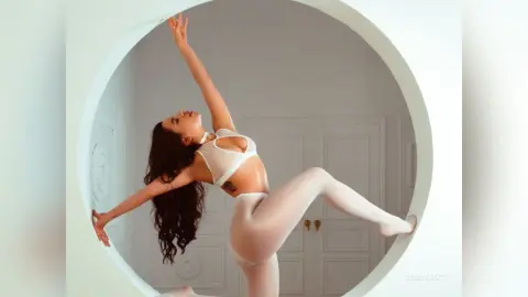 ElizaLuxe poses with her arms raised and one leg lifted, wearing a white bra and white leggings in a bright room.