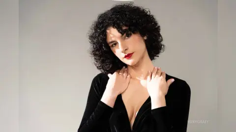 RoxyGraff poses with her hands on her neck, wearing a low-cut black long-sleeve top and red lipstick with her dark curly hair.