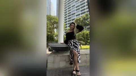 BiankaPerese poses outdoors in a black off-the-shoulder top and a long black and white patterned skirt with a high slit, standing next to a large white stone planter.