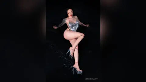 Tattooed woman in a silver bodysuit and high heels posing against a dark background with a glamorous mood.