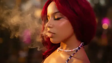 Young woman with red hair exhaling smoke in a warm, dreamy outdoor setting.
