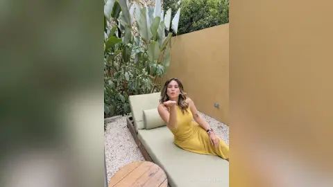 Smiling woman in a yellow summer dress relaxing on a lounge chair in a sunny tropical garden setting.