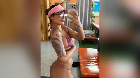 Woman in pink bikini and sunglasses taking a mirror selfie in a bright, modern room.