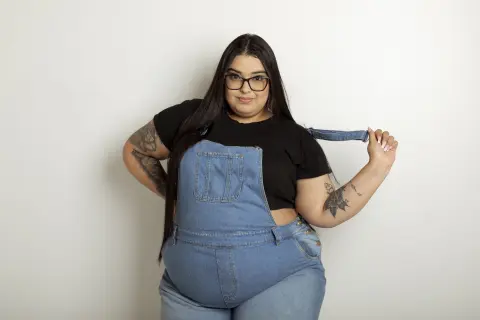 Curvy woman in denim overalls and black t-shirt smiling against a white background.