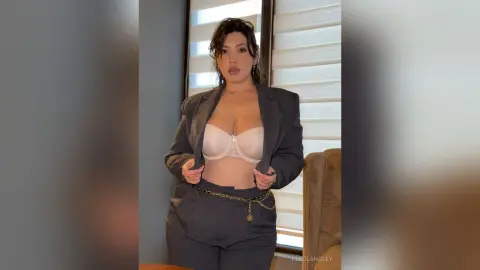 Confident woman in a grey blazer and white bralette posing in a sunlit room.