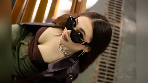 Stylish woman in a dark leather jacket and sunglasses posing with a chic, moody urban vibe.
