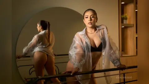 EvaCarusso poses in a sheer white button-down shirt and black bra, showing her cleavage and curves while leaning against a railing near a large mirror.
