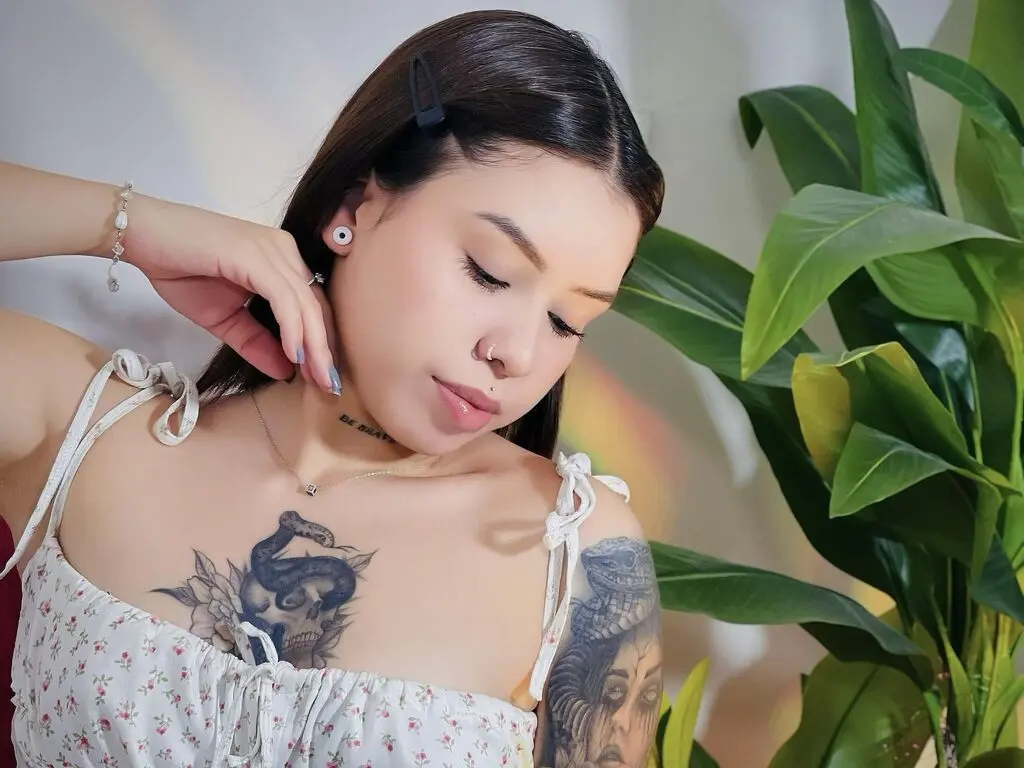 Young woman with tattoos in a white floral sundress posing near green houseplants in soft sunlight.