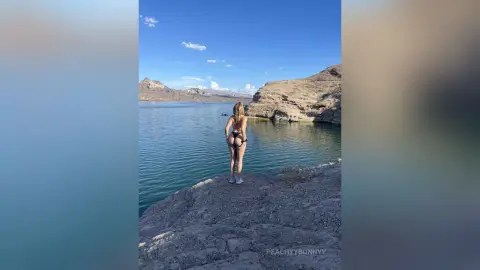 Blonde woman in a black bikini standing on a rocky cliff overlooking a serene blue lake under a clear sky.