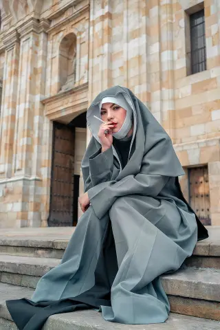Woman in a grey hooded nun habit sitting on stone steps outside a historic cathedral building.