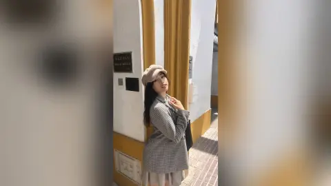 Stylish young woman in a grey plaid blazer and beige beret posing against a white building wall.