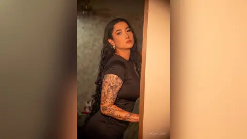 JuliahLewis poses against a wall wearing a tight black short-sleeve top that shows off her arm tattoos.