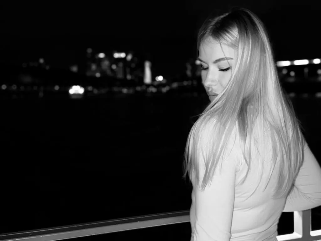 Blonde woman in a white long-sleeve shirt looking away against a dark city skyline at night.