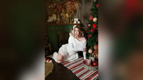 Young woman in a white dress sitting by a decorated Christmas tree in a cozy festive holiday setting.