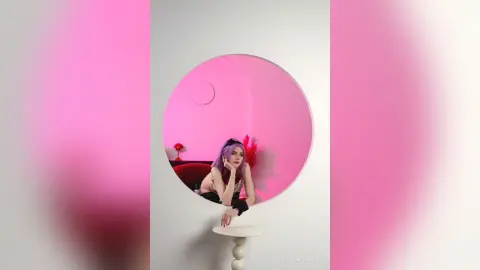 Young woman with purple hair in a black outfit sitting in a pink circular mirror setting with a dreamy mood.