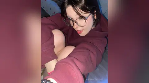 Young woman with glasses in a cozy maroon sweatshirt relaxing on a bed with a playful mood.