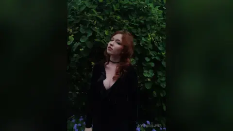 Redheaded woman in a black dress posing against a lush green leafy background with a moody aesthetic.