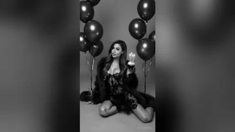 Glamorous woman in a black lace outfit and fur coat holding a birthday cake among floating balloons.