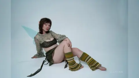 Young woman in green fairy costume with wings and knit leg warmers sitting on a white floor in a dreamy mood.