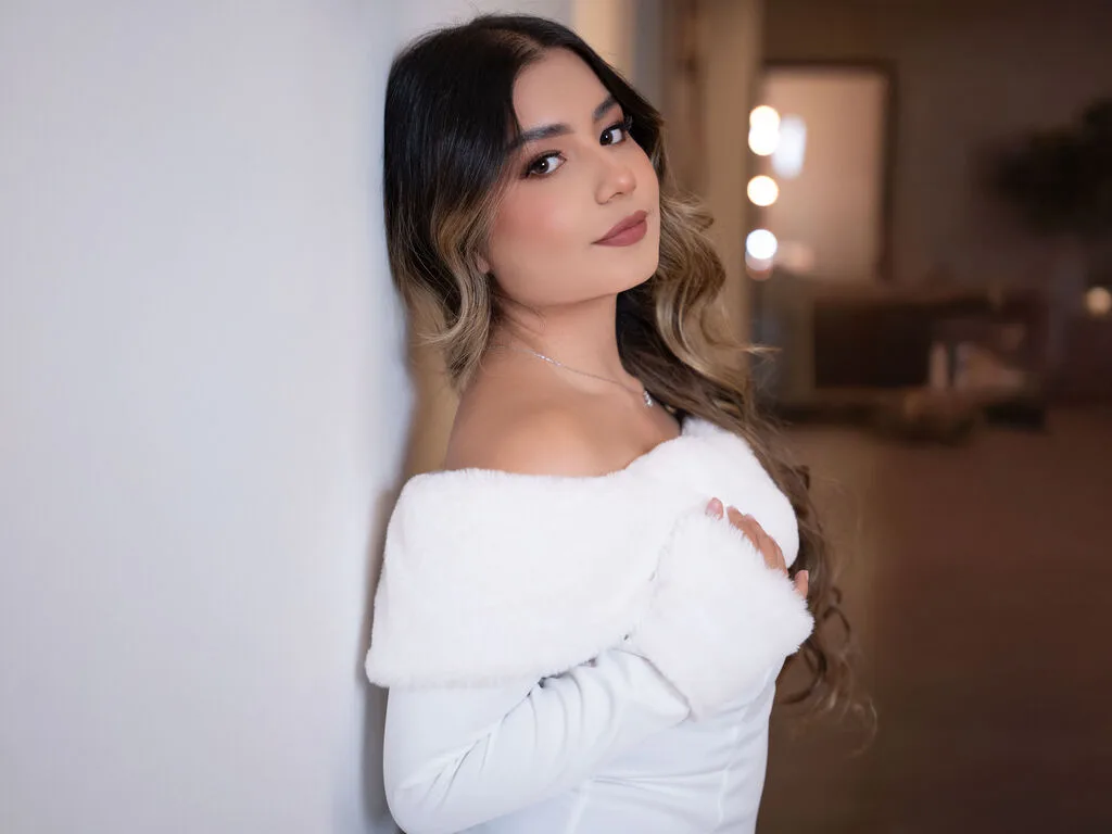 JessieOlivetti — JessieOlivetti poses against a white wall wearing a white off-the-shoulder furry top or shrug.