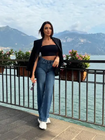 Stylish woman in black crop top and blue jeans posing by a lake with mountains in a serene summer setting.