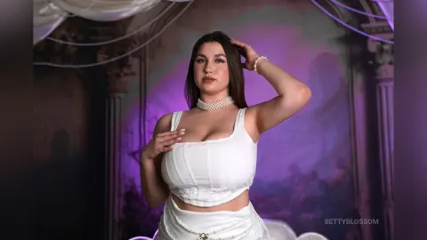 BettyBlossom — BettyBlossom poses in a white corset top and pearl necklace against a purple backdrop.