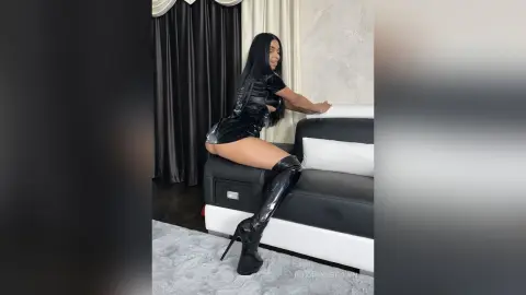 Glamorous woman in a black mini dress and thigh-high boots sitting on a modern sofa in a stylish room.