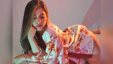 JessieLiu poses in a white floral print silk robe, looking down while resting her chin on her hand.