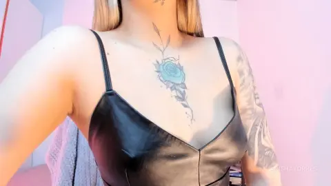 Woman in a black leather camisole top showing a large blue rose chest tattoo in soft pink lighting.