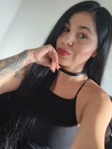 Smiling woman with long black hair and tattoos wearing a black camisole and choker in soft natural light.