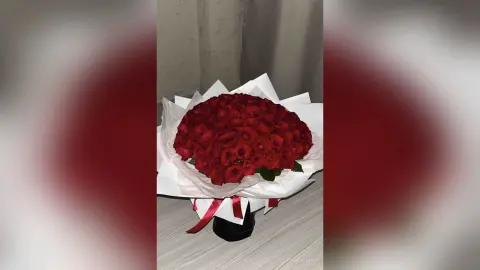 Large bouquet of deep red roses wrapped in white paper sitting on a light wooden table.