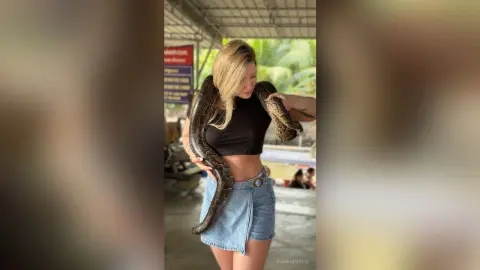 BiankaPerese poses outdoors holding a large brown snake around her shoulders and torso while wearing a black crop top and denim skirt.