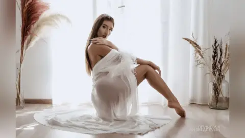 DeviNessa poses in a sheer white babydoll outfit, sitting on a white floor against a bright window background.