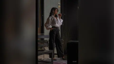 Elegant woman in white blouse and black trousers holding a glass in a moody, dimly lit modern interior.
