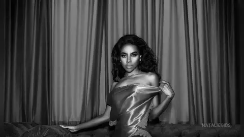 Elegant woman in a satin dress posing against draped curtains in a moody black and white portrait.