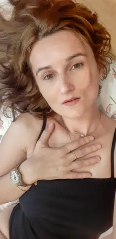 Redheaded woman in a black tank top lying on a floral bedsheet with a thoughtful expression.