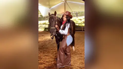 Cowgirl in brown leather chaps and cowboy hat standing with a dark horse in a rustic stable setting.
