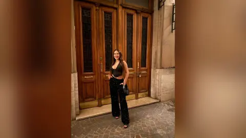 Smiling woman in a black bodysuit and patterned trousers standing before elegant vintage wooden doors.