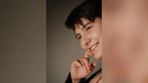 Smiling young man with dark hair posing in a casual black outfit against a neutral studio background.