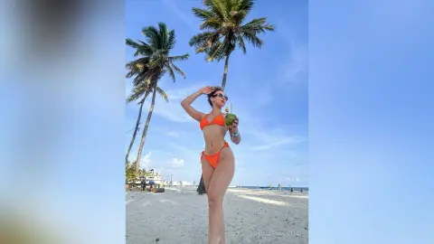 Woman in orange bikini holding a coconut on a sunny tropical beach with palm trees.