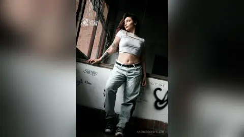 AnoraRockefeller poses in a white crop top and light blue jeans, showing her midriff and arm tattoo against a dark, urban background.