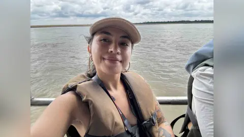 Smiling woman in a tan life vest enjoying a scenic boat ride on a calm river under a cloudy sky.