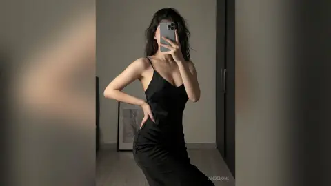 Elegant woman in a sleek black slip dress taking a mirror selfie in a modern minimalist room.