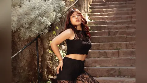 Young woman in a black lace crop top and skirt posing on stone stairs in a lush garden setting.
